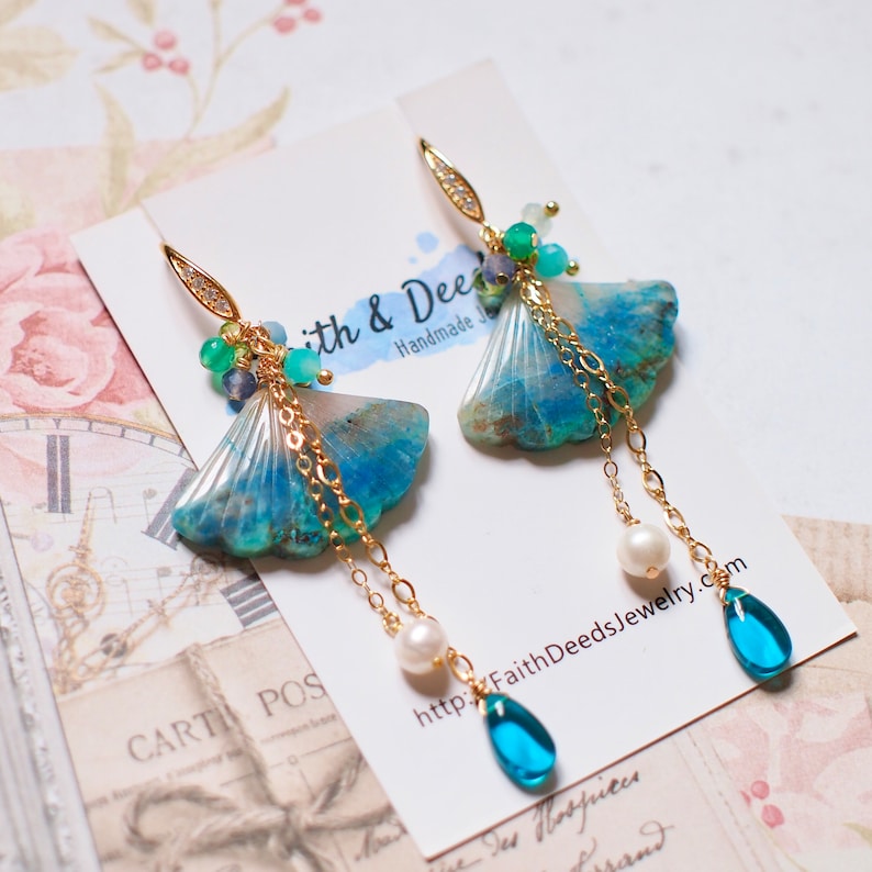 May include: A pair of long, dangling earrings featuring a blue and white shell-shaped charm, gold chains, and a variety of blue and green gemstones. The earrings are on a white card with the text "Faith & Deeds Handmade Jewelry" and a website address.