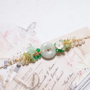 Könnte beinhalten: Ein zartes Armband mit einem runden Jade-Anhänger in Grün, umgeben von grünen und gelben Edelsteinen und einem weißen Blumen-Anhänger. Das Armband ist aus Golddraht gefertigt und hat eine zarte, feminine Ästhetik.