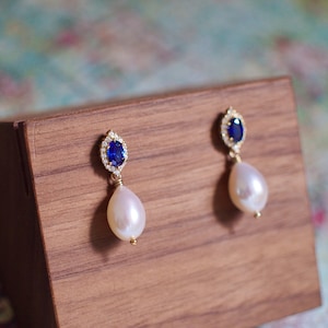 May include: A pair of gold earrings with a blue sapphire gemstone surrounded by smaller clear gemstones and a white pearl drop.