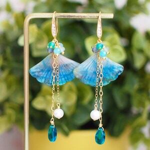 May include: A pair of gold dangle earrings featuring blue and green gemstone accents, a shell-shaped pendant, and a pearl drop.