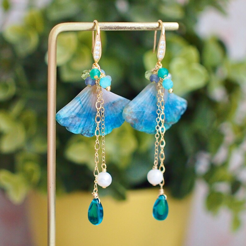 May include: A pair of blue and green gemstone earrings with gold chains and a pearl accent. The earrings are shaped like a fan and have a teardrop-shaped blue gemstone dangling from the bottom.