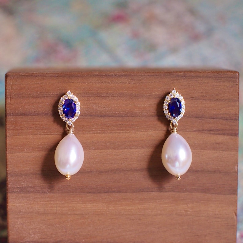 May include: A pair of gold earrings with a blue sapphire gemstone surrounded by a halo of clear cubic zirconia stones. Each earring features a white pearl drop.