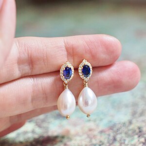 May include: Gold earrings with a blue sapphire gemstone surrounded by clear cubic zirconia stones and a white pearl drop.