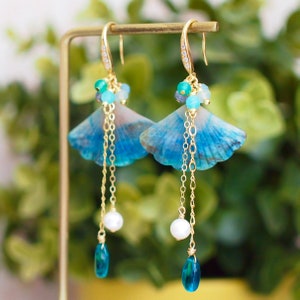 May include: A pair of blue and gold earrings with a shell-shaped design. The earrings feature a long chain with a pearl and a blue teardrop-shaped stone.