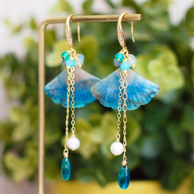 May include: A pair of blue and gold earrings with a shell-shaped design. The earrings feature a long chain with a pearl and a blue teardrop-shaped stone.