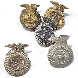 May include: A collection of five circular metal pins featuring an eagle emblem at the top. Each pin displays the text FFA, Vocational Agriculture, and a central sunburst design. The pins vary in metallic finishes, including gold, silver, and bronze tones.