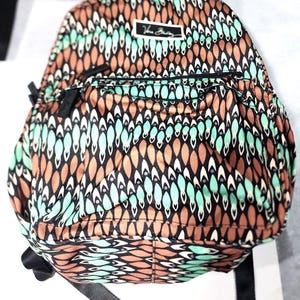 May include: A Vera Bradley backpack featuring a repeating pattern of brown, mint green, and black teardrop shapes. The bag includes a front zippered pocket and a rectangular brand label with the text Vera Bradley centered at the top.