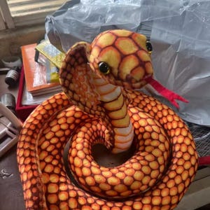 May include: A plush cobra toy featuring a vibrant orange and yellow scale pattern across its coiled body. The snake has a raised hood, large black eyes, and a bright red forked tongue protruding from its mouth, with a light cream-colored underbelly.