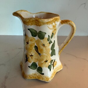 May include: A ceramic pitcher with a cream-colored base, featuring a hand-painted floral pattern of yellow blossoms and green leaves. The rim and base have a textured, golden-yellow sponge-painted border, and the pitcher has a curved handle.