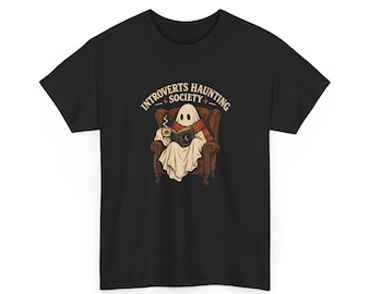 Peut inclure: Un t-shirt noir avec un motif représentant un fantôme blanc portant une écharpe rouge, assis dans un fauteuil marron, lisant un livre et tenant une tasse fumante. Le texte au-dessus du fantôme indique INTROVERTS HAUNTING SOCIETY en beige.