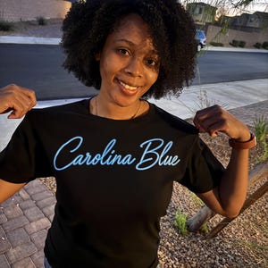 May include: A black short-sleeved t-shirt featuring the text "Carolina Blue" printed in a light blue script font across the chest. The shirt has a crew neckline and a standard fit, showcasing the graphic design clearly against the dark fabric.