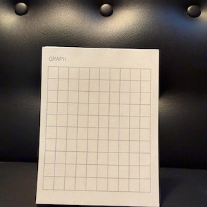 May include: A rectangular white paper notepad featuring a grid of thin black lines. The word GRAPH is printed in black capital letters in the top left corner of the page. The notepad consists of multiple sheets of paper stacked together.