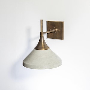May include: A wall-mounted light fixture with a concrete shade and brass accents. The fixture has a square backplate and a conical shade. The brand name "CORDERO HOME & LIGHTING" is in the upper right corner.
