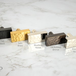 Könnte beinhalten: Fünf rechteckige Schrankknöpfe in verschiedenen Ausführungen: mattschwarz, satiniertes Messing, gebürstetes Nickel, geöltes Bronze und poliertes Nickel. Die Knöpfe haben eine gehämmerte Textur.