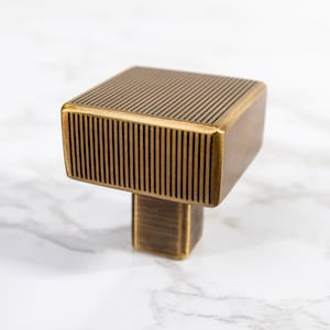 May include: A square, bronze-colored cabinet knob with a ribbed top and a rectangular base. The knob is a warm, metallic tone, and the design is modern and geometric. The knob is likely for a drawer or cabinet. The brand name "CORDERO" is visible.
