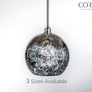 Könnte beinhalten: Eine silberne Pendelleuchte mit einem schwarzen und weißen, gesprenkelten Glasglobus-Schirm. Die Leuchte hängt an einem silbernen Kabel. Der Text "CORDERO HANDCRAFTED LIGHTING" ist oben im Bild sichtbar. Der Text "3 Sizes Available" ist unten im Bild sichtbar.