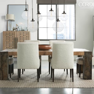 May include: A rustic wooden dining table with six white upholstered chairs and a copper bowl in the center. The table has a distressed finish and metal legs. The chairs have a fluffy, white fabric covering. The table is set in a modern dining room with large windows and a white wall.
