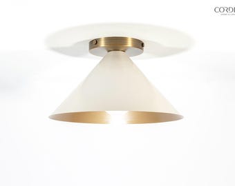 May include: A modern ceiling light fixture with a white conical shade and a gold-colored interior. The fixture has a brass-colored circular base. The light is turned on, illuminating the gold interior. The brand name "CORDERO" is visible.