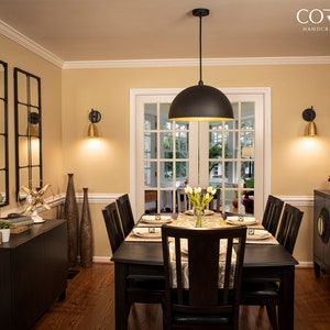 May include: A black pendant light with a gold interior hangs over a dining table with six chairs. The table is set with white plates, gold silverware, and a vase of white flowers. The room is decorated with gold accents and mirrors.