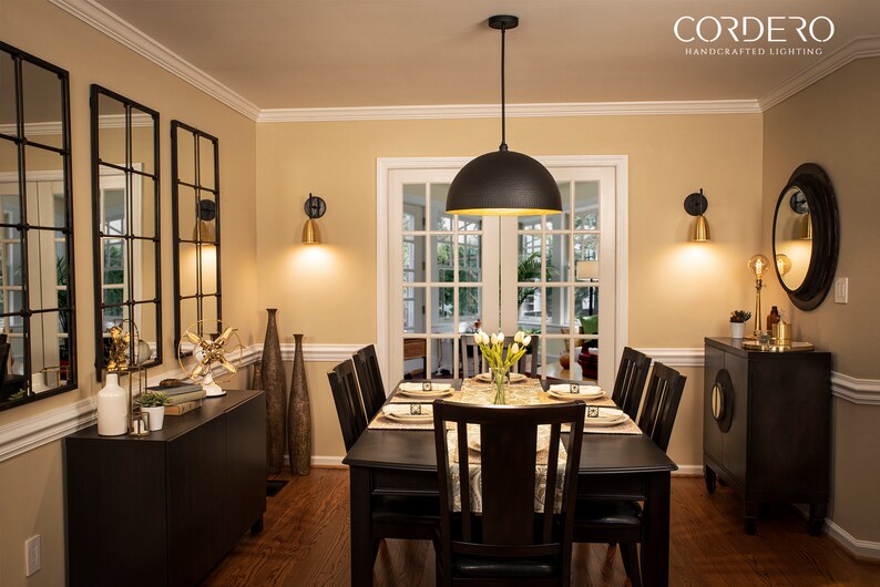 May include: A black pendant light with a gold interior hangs over a dining table with six chairs. The table is set with white plates, gold silverware, and a vase of white flowers. The room is decorated with gold accents and mirrors.