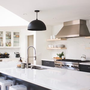 May include: A modern kitchen with white cabinets, a white countertop, and a black pendant light. The kitchen features a stainless steel range hood, a farmhouse sink, and a white quartz countertop.