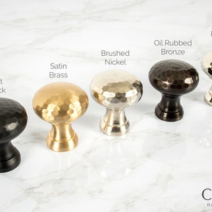 May include: A collection of five hammered metal cabinet knobs in various finishes: Flat Black, Satin Brass, Brushed Nickel, Oil Rubbed Bronze, and Polished Nickel. The knobs have a rounded, textured design and are displayed on a marble surface.