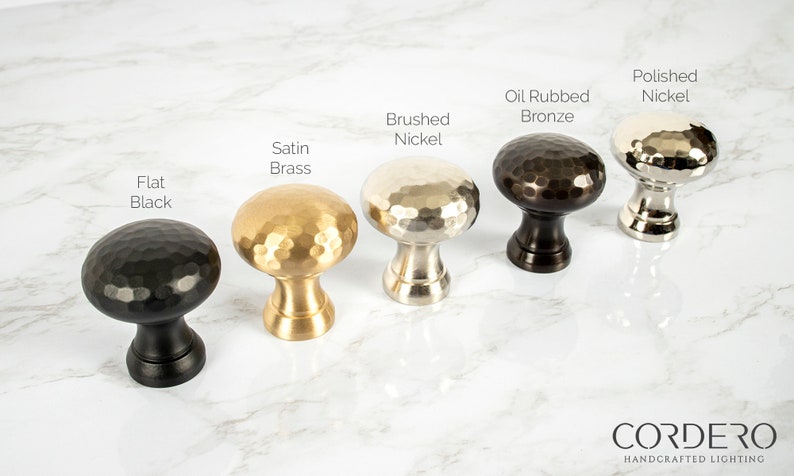 May include: A collection of five hammered metal cabinet knobs in various finishes: Flat Black, Satin Brass, Brushed Nickel, Oil Rubbed Bronze, and Polished Nickel. The knobs have a rounded, textured design and are displayed on a marble surface.