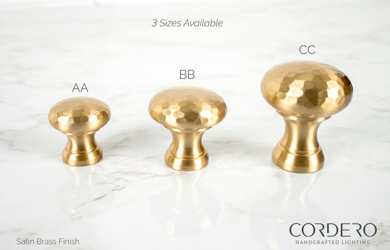 May include: Three satin brass finish cabinet knobs in varying sizes, labeled AA, BB, and CC. Each knob features a hammered texture on the rounded top and a cylindrical base. The text "3 Sizes Available" is above the knobs, and the Cordero Handcrafted Lighting logo is at the bottom.