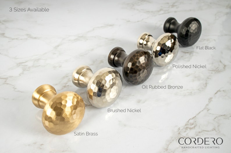 May include: A selection of decorative cabinet knobs in various finishes, including satin brass, brushed nickel, oil-rubbed bronze, polished nickel, and flat black. The knobs have a textured, hammered design. The text "3 Sizes Available" is visible.