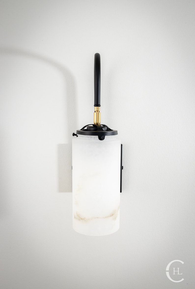 May include: A black wall sconce with a white alabaster glass shade. The sconce has a curved arm and a brass accent.