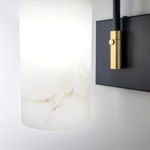 May include: A white alabaster wall sconce with a black metal base and a gold metal switch. The sconce is mounted on a white wall.