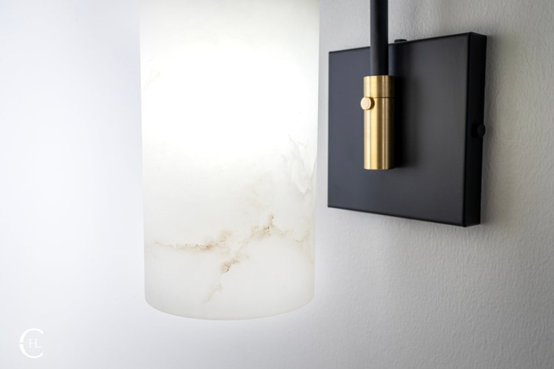 May include: A white alabaster wall sconce with a black metal base and a gold metal switch. The sconce is mounted on a white wall.