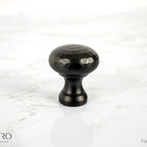 May include: A black cabinet knob with a hammered texture on the rounded top. The knob has a flat black finish and a cylindrical base. The image also includes the text "CORDERO HANDCRAFTED LIGHTING" and "Flat Black Finish".