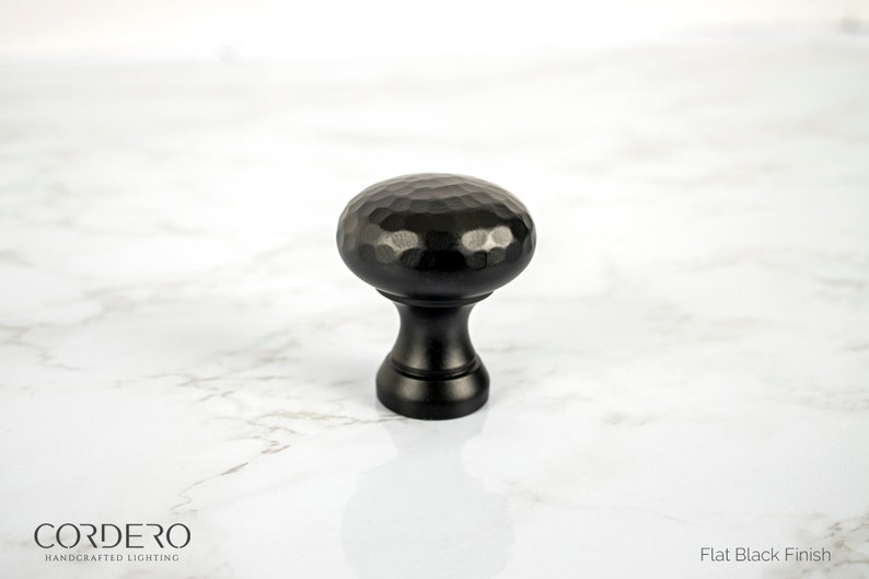 May include: A black cabinet knob with a hammered texture on the rounded top. The knob has a flat black finish and a cylindrical base. The image also includes the text "CORDERO HANDCRAFTED LIGHTING" and "Flat Black Finish".