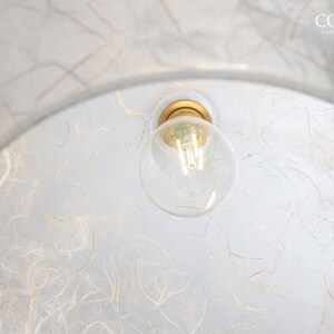May include: A white, round, paper lampshade with a gold-colored light fixture and a clear light bulb. The lampshade has a textured surface with thin, gold-colored lines.
