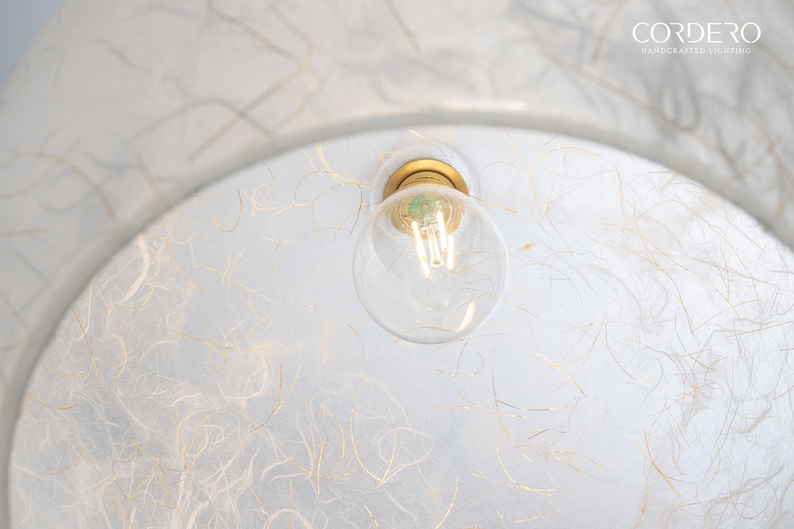 May include: A white, round, paper lampshade with a gold-colored light fixture and a clear light bulb. The lampshade has a textured surface with thin, gold-colored lines.