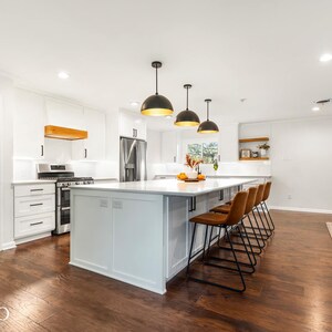 May include: A white kitchen with a large island featuring a white countertop and black bar stools. The island has a white cabinet with a built-in shelf. There are three black pendant lights hanging over the island. The kitchen has white cabinets and a stainless steel refrigerator.