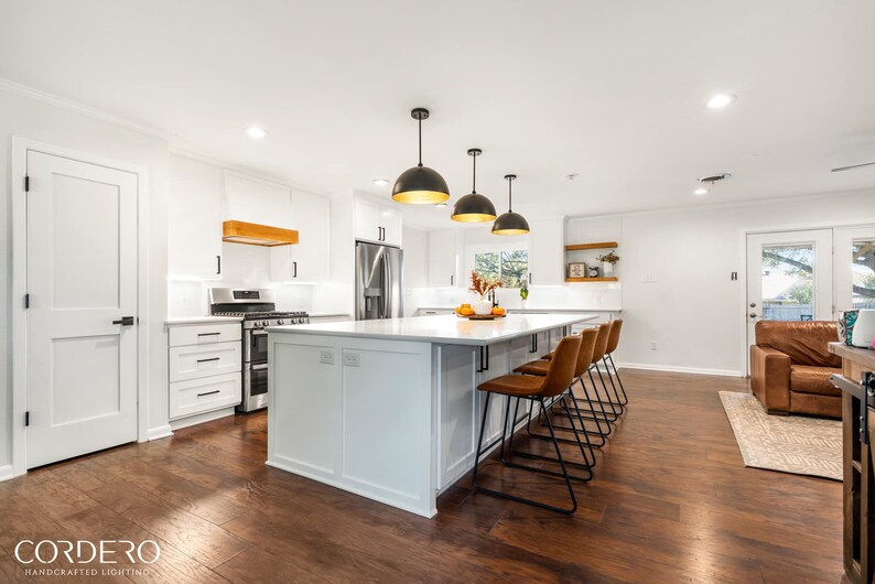 May include: A white kitchen with a large island featuring a white countertop and black bar stools. The island has a white cabinet with a built-in shelf. There are three black pendant lights hanging over the island. The kitchen has white cabinets and a stainless steel refrigerator.