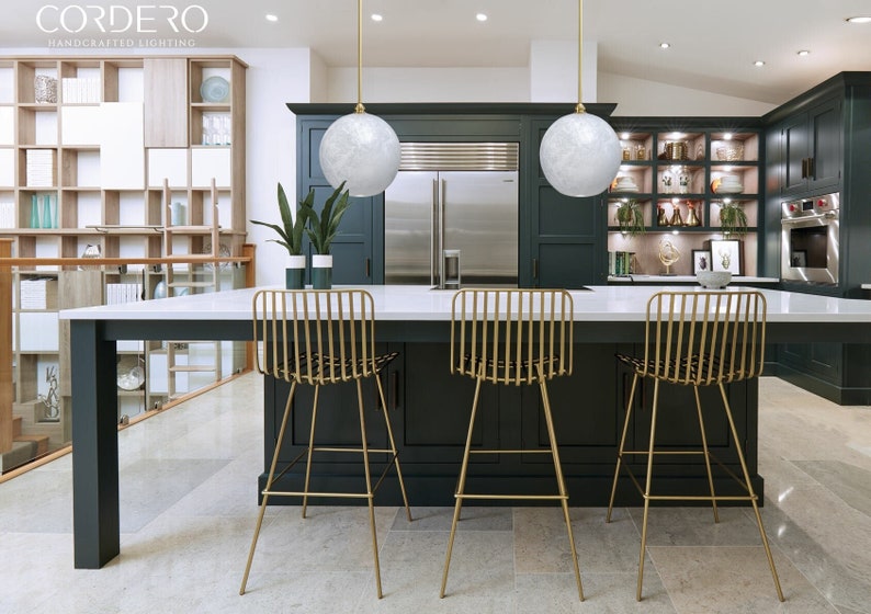 May include: Three gold metal bar stools with a woven seat back and a white countertop in a kitchen with dark green cabinets and two white globe pendant lights.