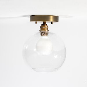 May include: A modern ceiling light fixture with a clear, textured glass globe shade. The fixture has a brass-colored base and a visible light bulb. The design is minimalist and suitable for various interior styles.