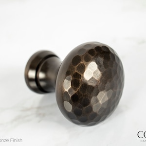 May include: Oil rubbed bronze finish cabinet knob with a hammered texture. The hardware has a dark brown color and a round shape. The product is from Cordero Handcrafted Lighting.