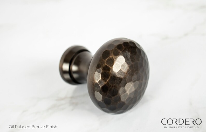 May include: Oil rubbed bronze finish cabinet knob with a hammered texture. The hardware has a dark brown color and a round shape. The product is from Cordero Handcrafted Lighting.