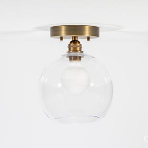 May include: A modern ceiling light fixture with a clear, spherical glass shade. The fixture has a brass-colored base and a visible light bulb. The design is minimalist and suitable for various interior styles.