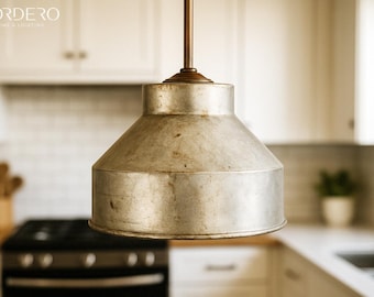 May include: A silver metal pendant light fixture with a wide, rounded shade hangs from a ceiling in a kitchen. The light fixture is in focus, while the kitchen counter and cabinets are blurred in the background.