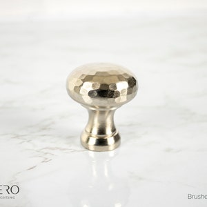 May include: A brushed nickel finish cabinet knob with a hammered texture on the rounded top. The knob has a short, cylindrical base and is designed for furniture or cabinetry. The image also includes the text "CORDERO HANDCRAFTED LIGHTING" and "Brushed Nickel Finish".