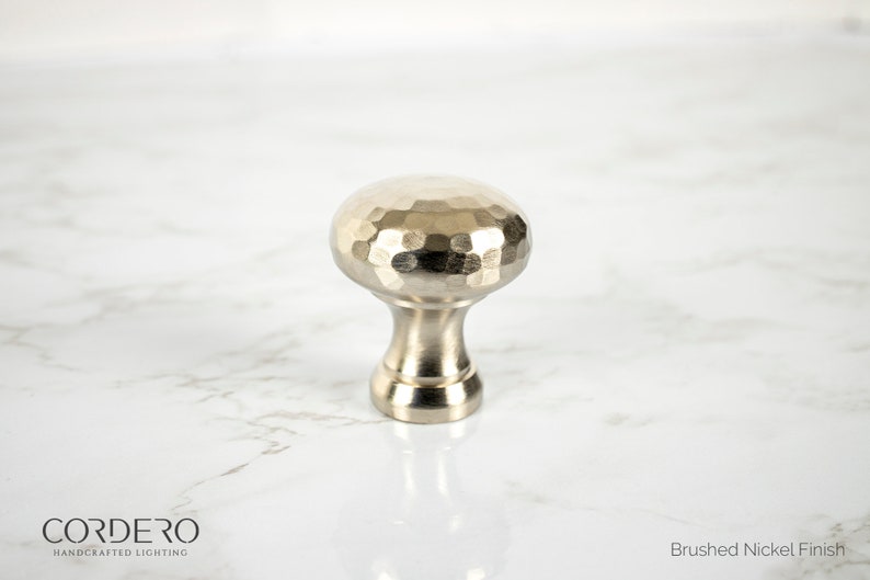 May include: A brushed nickel finish cabinet knob with a hammered texture on the rounded top. The knob has a short, cylindrical base and is designed for furniture or cabinetry. The image also includes the text "CORDERO HANDCRAFTED LIGHTING" and "Brushed Nickel Finish".