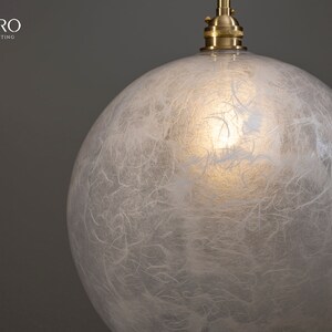 May include: A white, round, glass pendant light fixture with a textured, paper-like surface. The light fixture is suspended from a brass cord.