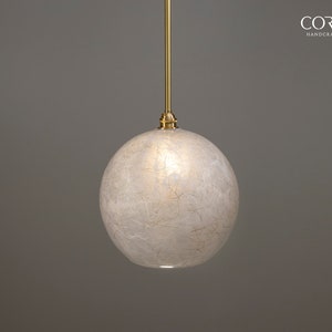 May include: A white globe pendant light with a brass cord and a brass ceiling fixture. The globe is made of a translucent material with a textured surface. The light is hanging from a gray ceiling.