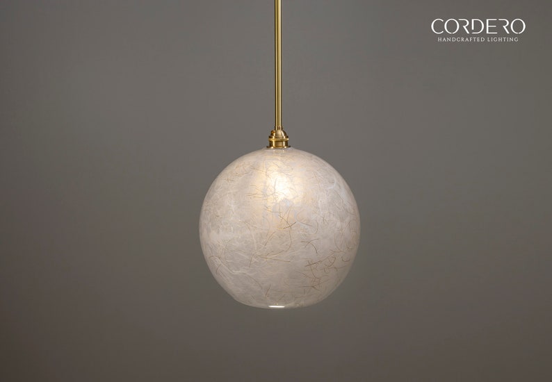 May include: A white globe pendant light with a brass cord and a brass ceiling fixture. The globe is made of a translucent material with a textured surface. The light is hanging from a gray ceiling.
