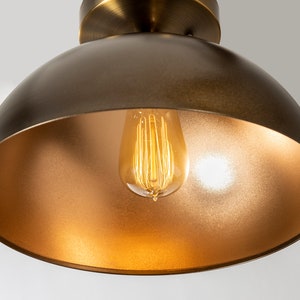 May include: A gold-toned metal dome pendant light fixture with a clear glass light bulb inside. The light is on and the bulb is glowing.